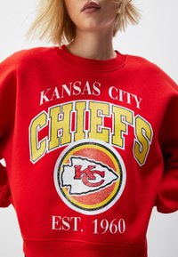 Sudadera roja con "Kansas City Chiefs" en texto amarillo, un logo circular en el centro y "EST. 1960" en blanco en la parte inferior. Material de algodón.