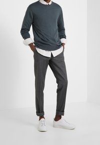 Maglione grigio a maniche lunghe indossato sopra una camicia bianca con colletto, abbinato a pantaloni grigi arrotolati e sneaker bianche. Tessuto liscio, vestibilità casual.
