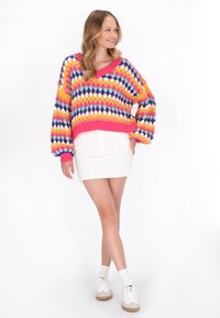 Kolorowy, oversized sweter z dekoltem w serek, ozdobiony geometrycznym wzorem w żywych kolorach, zestawiony z białą spódnicą i trampkami.