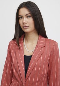 Rust-colored blazer with white pinstripes, structured design, notched collar, and soft texture. Features a silver chain necklace.