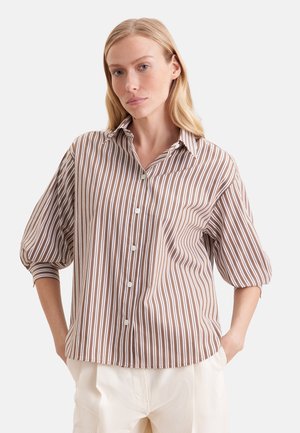 Kobieta w brązowo-białej koszuli w pionowe paski z bufkami na rękawach 3/4 i kremowych spodniach, stoi z rękami w kieszeniach.