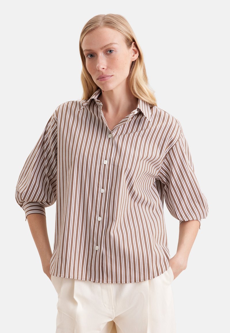 Femme portant une chemise boutonnée rayée verticalement marron et blanche avec des manches ballon 3/4 et un pantalon de couleur crème, debout les mains dans les poches.