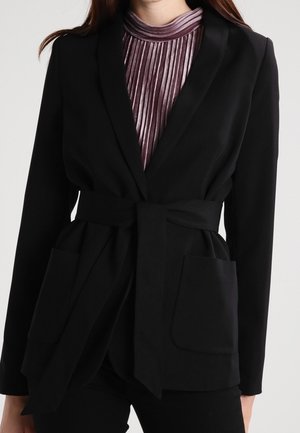 Vrouw draagt een zwarte blazer met riem en grote zakken over een geplooide, glanzende paarse top met hoge hals.