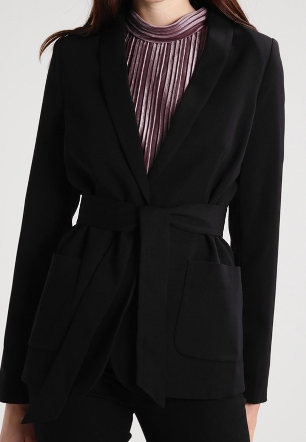 Femme portant un blazer noir ceinturé avec de grandes poches sur un top plissé, brillant et violet à col montant.