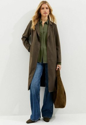 Vrouw in een lange bruine jas, olijfgroene blouse, wijde blauwe jeans, met een grote bruine suède tas, staand tegen een effen witte muur.