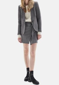 Mujer con un blazer texturizado gris y una falda ceñida a juego con cinturón, un suéter de punto color crema y botas de tobillo negras con tachuelas y cordones.