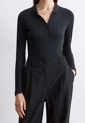 Frau trägt ein tailliertes schwarzes, geripptes Langarmhemd mit Kragen und Knöpfen sowie locker sitzende schwarze High-Waist-Hosen, eine Hand in der Tasche.