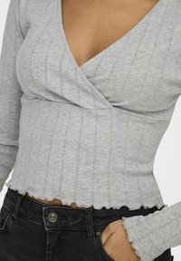 Grijze lange mouwen top met een V-hals, voorzien van een geribbelde textuur en open stikpatronen, eindigend met een gegolfde zoom. Gepaard met zwarte jeans.