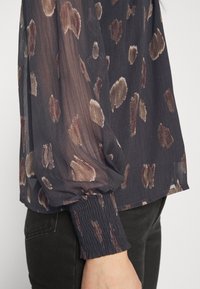 Blusa trasparente di un blu navy scuro con motivi astratti marroni e beige, caratterizzata da polsini arricciati e una silhouette fluida.