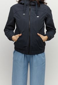 Granatowa kurtka przeciwwiatrowa z zamkiem z przodu, regulowanym kapturem, elastycznymi mankietami oraz bocznymi kieszeniami. Gładki materiał z dopasowaną talią.