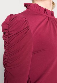 Burgundin pitkähihaisia paita, jossa on rypistettyä, teksturoitua kangasta olkapäillä ja kauluksessa. Sujuva materiaali, jossa on hieman kiiltävä pinta.