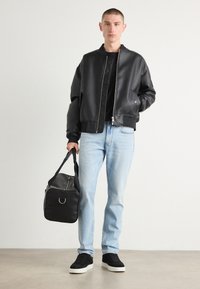 Schwarze Kunstleder-Bomberjacke mit Reißverschluss, graues Rundhals T-Shirt, hellblaue Jeans und schwarze Tasche mit silberner Akzentuierung. Schwarze Sneakers.