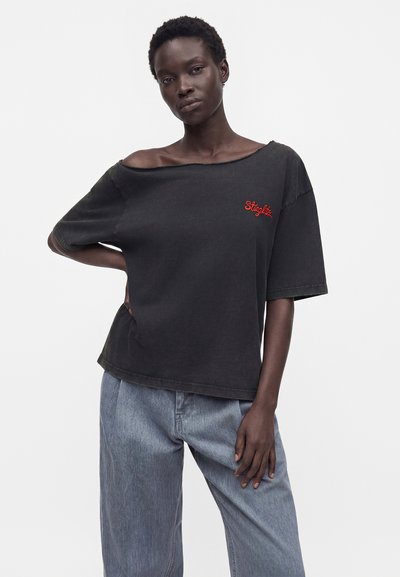 Γυναίκα που φοράει μαύρο φαρδύ μπλουζάκι off-shoulder με κόκκινο κείμενο "Stugltz" και ανοιχτό μπλε τζιν με φαρδιά πόδια, σε απλό λευκό φόντο.