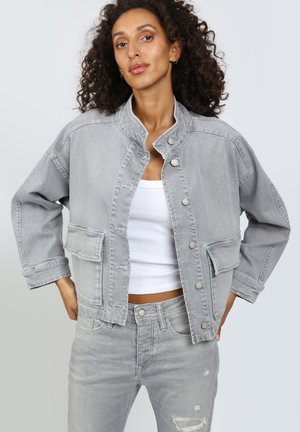 Frau mit lockigem Haar, die eine hellgraue Jeansjacke über einem weißen, bauchfreien Tanktop und passenden hellgrauen, abgenutzten Jeans trägt.