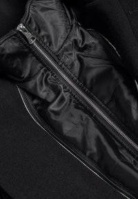 Musta takki, jossa on vetoketjullinen nylonvuori, sileä pinta ja kontrastiväriset tikkaukset reunassa.