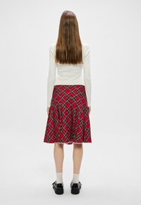 Jupe à carreaux rouges avec des accents verts et blancs, design plissé. Portée avec un haut à manches longues blanc et des chaussures noires, vue de l'arrière.