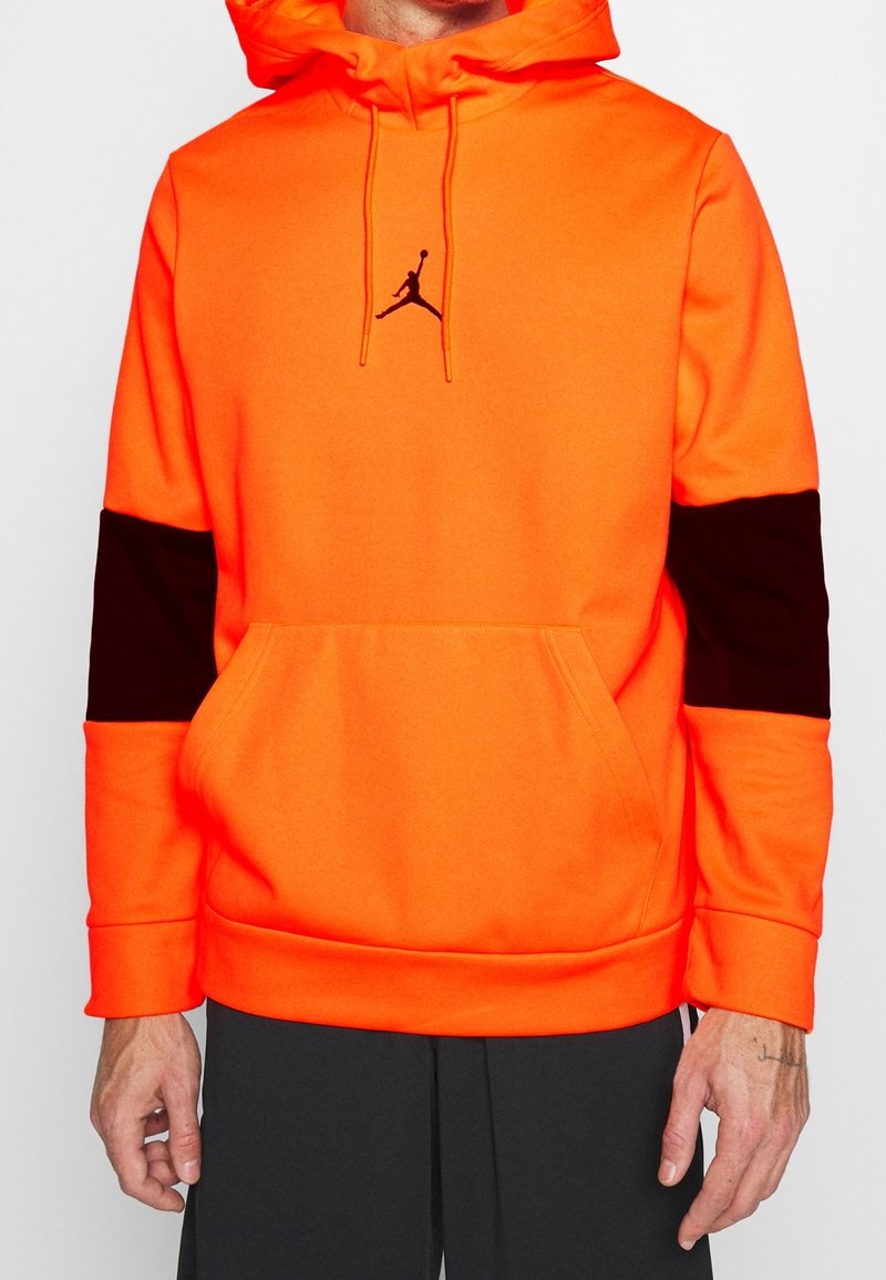 Sweat à capuche orange vif avec des patches noirs sur les manches, une poche avant et un logo Jumpman noir centré sur la poitrine, porté avec un pantalon noir.