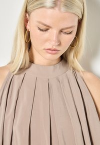 Mujer rubia con el cabello lacio lleva pendientes de aro dorados torcidos y una blusa sin mangas taupe plisada de cuello alto, mirando hacia abajo.