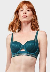 Reggiseno sottile color teal con rete testurizzata, bordi scallop e una fascia a righe orizzontali. Presenta spalline sottili regolabili e un design con apertura.