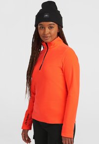 Suéter de polar naranja brillante con media cremallera en el frente, cuello alto y acentos en negro. Presenta el logo "Polartec" en la manga.