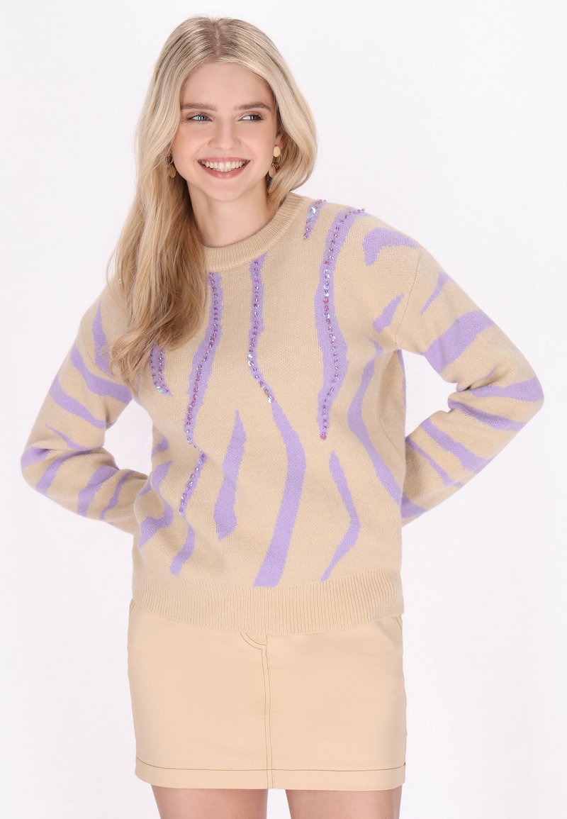 Beige Pullover mit lila Streifen und Pailletten. Gerippter Halsausschnitt und Bündchen. Schmal geschnittenes Design. Kombiniert mit einem hellbeigen Minirock.
