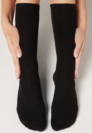 Svarta ribbade knähöga strumpor med en texturerad yta, som visas två strumpor hållna vertikalt med händer på varje sida.