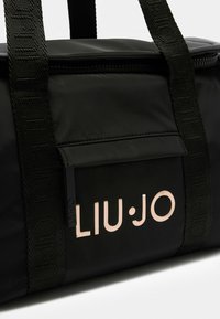 Sac de sport noir avec logo en rose gold, tissu lisse, fermeture éclair et sangles tissées avec texte. Design compact et fonctionnel.