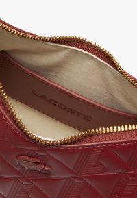 Sac rouge texturé avec un motif géométrique, fermeture éclair dorée et doublure intérieure beige. Logo Lacoste embossé à l'intérieur.