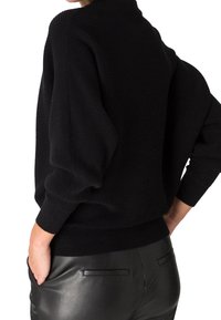 Personne portant un pull côtelé noir à manches ballon et un pantalon en cuir noir, vue de dos avec les mains dans les poches.