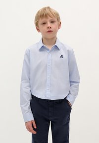 Camicia button-up a righe blu chiaro con un piccolo emblema di teschio navy, abbinata a pantaloni navy; presenta un colletto classico e maniche lunghe.
