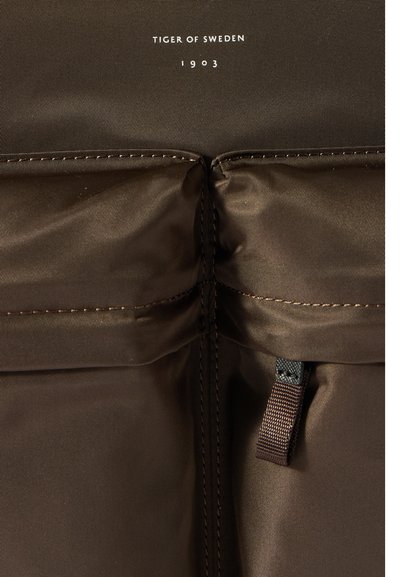 Ruda, tekstūruota medžiaga su matomu siuvimu ir užtrauktuku. Prekės ženklas skaito "TIGER OF SWEDEN 1903" šalia viršaus.