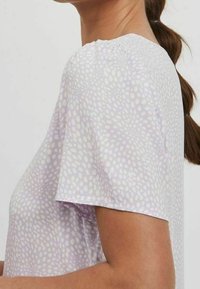 Žena v světle fialovém topu s krátkými rukávy, pokrytém nepravidelným bílým tečkovaným vzorem, s vlasy staženými do culíku.