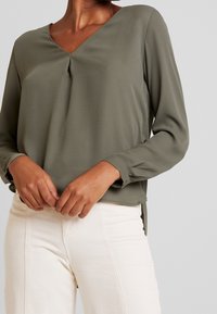 Olivgrünes, langärmliges Blusenshirt mit V-Ausschnitt, das eine Faltenoptik vorne und einen geraden Saum hat, kombiniert mit hellen Hosen.