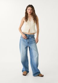 Beige linne kort topp med knapprad; högmidjade, löst sittande ljusblå jeans med blek finish; bruna platåsandaler.