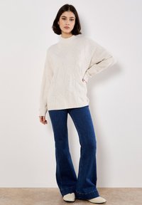Jasny kremowy sweter z teksturowanymi, diamentowymi wzorami; prążkowany okrągły dekolt i mankiety. Połączony z dzwonowymi niebieskimi dżinsami i jasnymi butami.