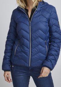 Giacca a vento imbottita blu navy con cuciture a chevron, zip frontale e cappuccio regolabile. Presenta due tasche laterali e polsini a costine.