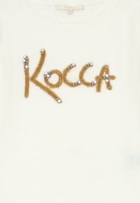 T-shirt in cotone crema con ricamo "Kocca" in filato marrone, adornato con perline trasparenti e metalliche che valorizzano il design del testo.