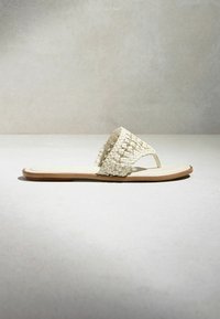 Sandale plate avec large bride tressée sur les orteils en cuir écru et semelle marron, présentée de profil sur fond neutre.