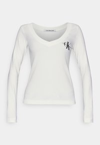 Vit långärmad T-shirt med V-hals i ribbat tyg. Har en svart "CK"-logotyp på vänster bröst. Enkel design, inga mönster.