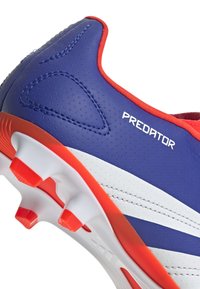 Zapato de fútbol azul con acentos rojos, que presenta una parte superior sintética texturizada, costuras prominentes y rayas blancas a lo largo de los lados.