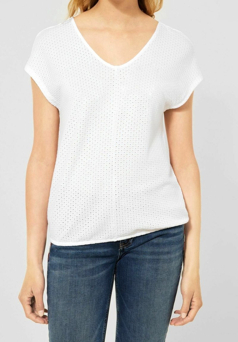 Haut blanc à manches courtes avec un col en V, présentant un motif texturé de petits trous. Associé à un jean bleu foncé.