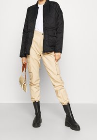 Svart vadderad jacka, beige cargobyxor med sidofickor, svarta anklarboots och en liten texturerad väska med handtag i brunt.