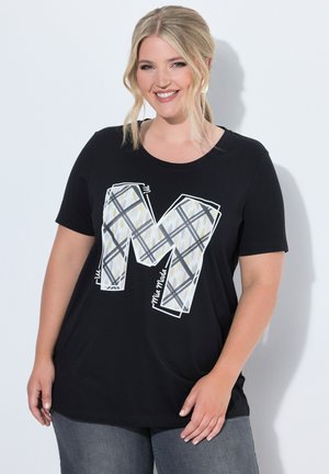 Donna sorridente con capelli biondi indossa una maglietta nera a maniche corte con una grande lettera M decorata e pantaloni grigi casual.