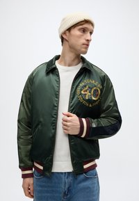 Πράσινο σατέν μπολερό τζάκετ με κεντημένες λεπτομέρειες, κλείσιμο με φερμουάρ, ριμπ μανσέτες και γιακά. Διαθέτει ριγέ διακοσμητικά σε μπορντό και μπεζ χρώμα.