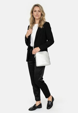 Woman with blonde hair wears black blazer, white shirt, black pants, black loafers, and holds a silver crossbody bag against a white background.