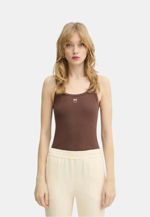 Donna bionda con capelli mossi indossa un top marrone senza maniche con piccolo logo bianco e pantaloni beige chiaro, rivolta in avanti su sfondo bianco.