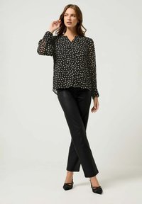 Blusa nera trasparente con leggeri pois, scollatura a V e maniche lunghe. Abbinata a pantaloni neri aderenti e scarpe con tacco nere.