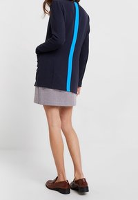 Námořnický cardigan s dlouhými rukávy, který má na zadní straně jasně modrý vertikální pruh. Nositý přes kostkovanou sukni a hnědé boty.