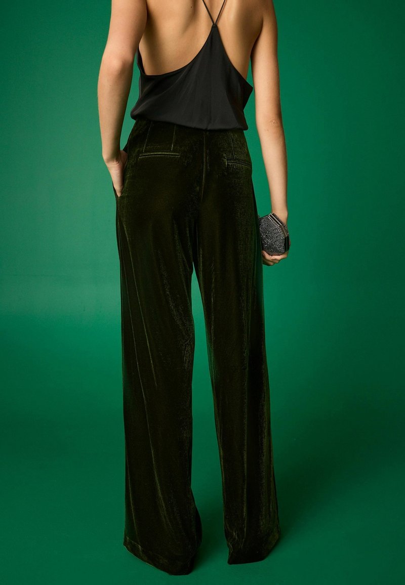 Femme portant un haut dos nu noir et un pantalon large en velours vert foncé, tenant une petite pochette noire sur fond vert.