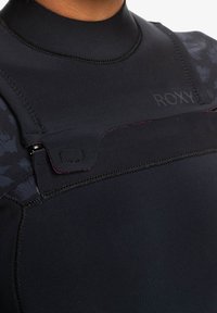 Black neoprene wetsuit with a high neck, featuring a central zipper and the "ROXY" logo embossed on the chest area. Subtle floral pattern on the side.